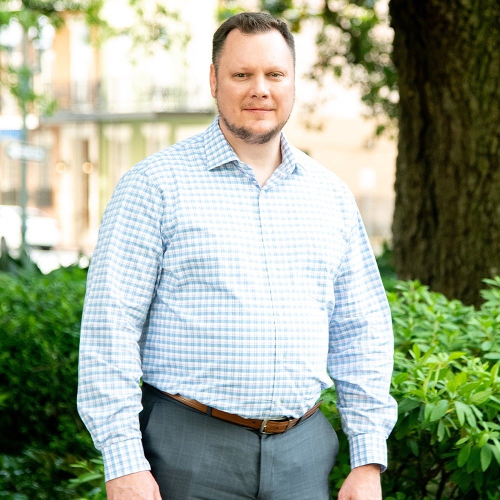 A man in a gray shirt demonstrating leadership as he stands confidently in front of bushes.