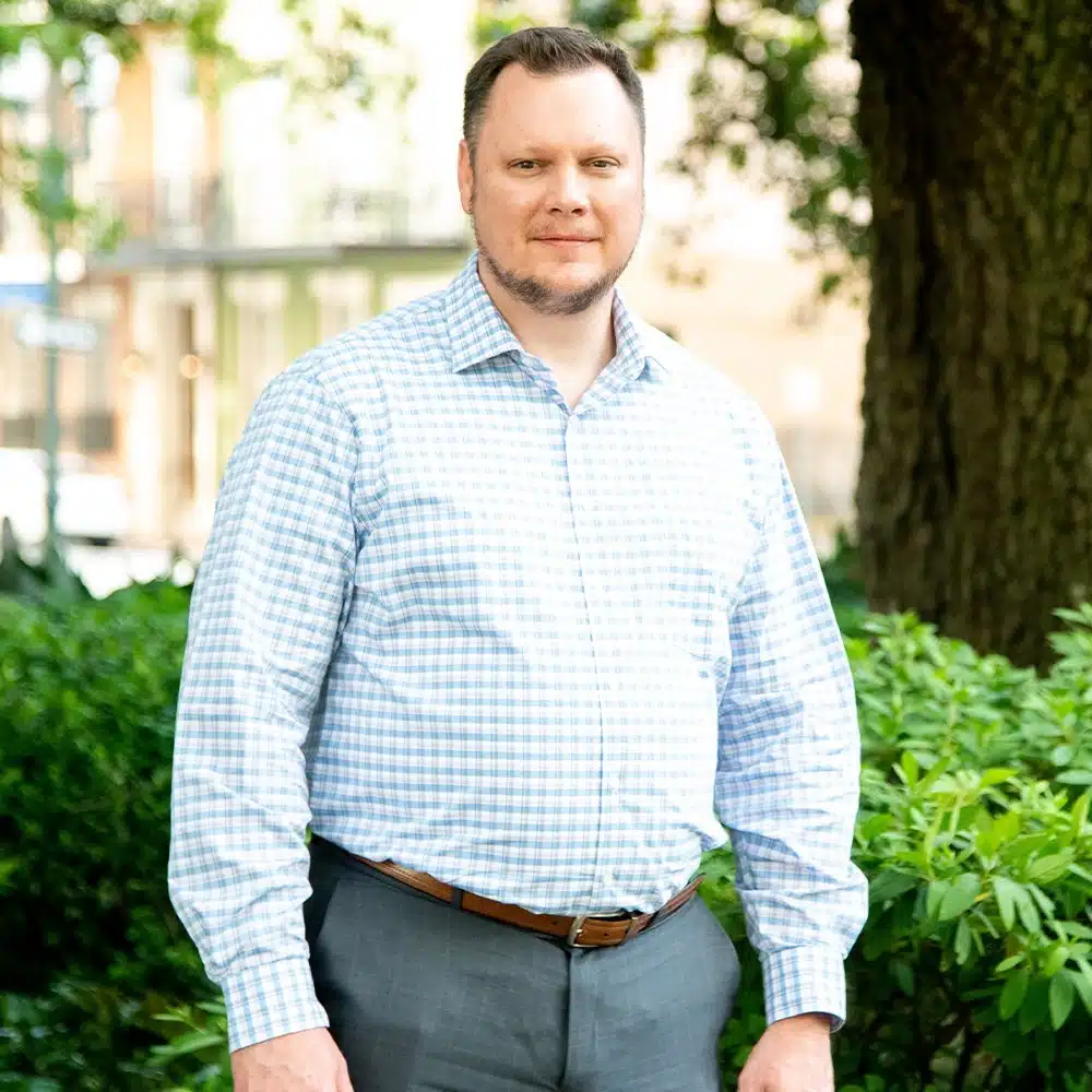 A man in a gray shirt demonstrating leadership as he stands confidently in front of bushes.