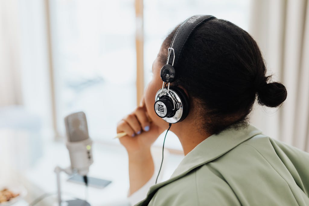 A woman wearing headphones is engaged in podcasting indoors by a window.